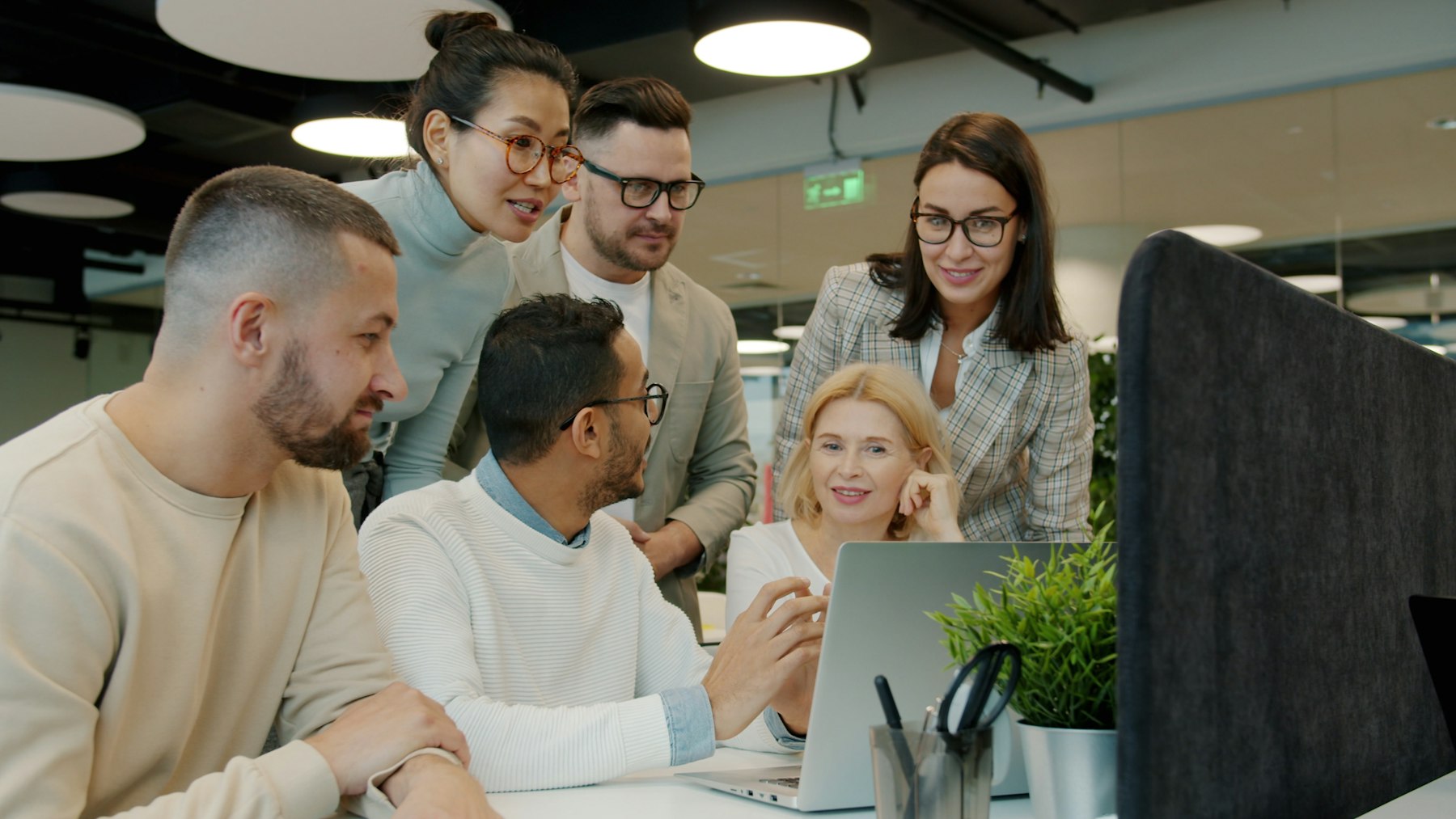 A diverse group of colleagues gathered around a laptop discussing what's on screen.